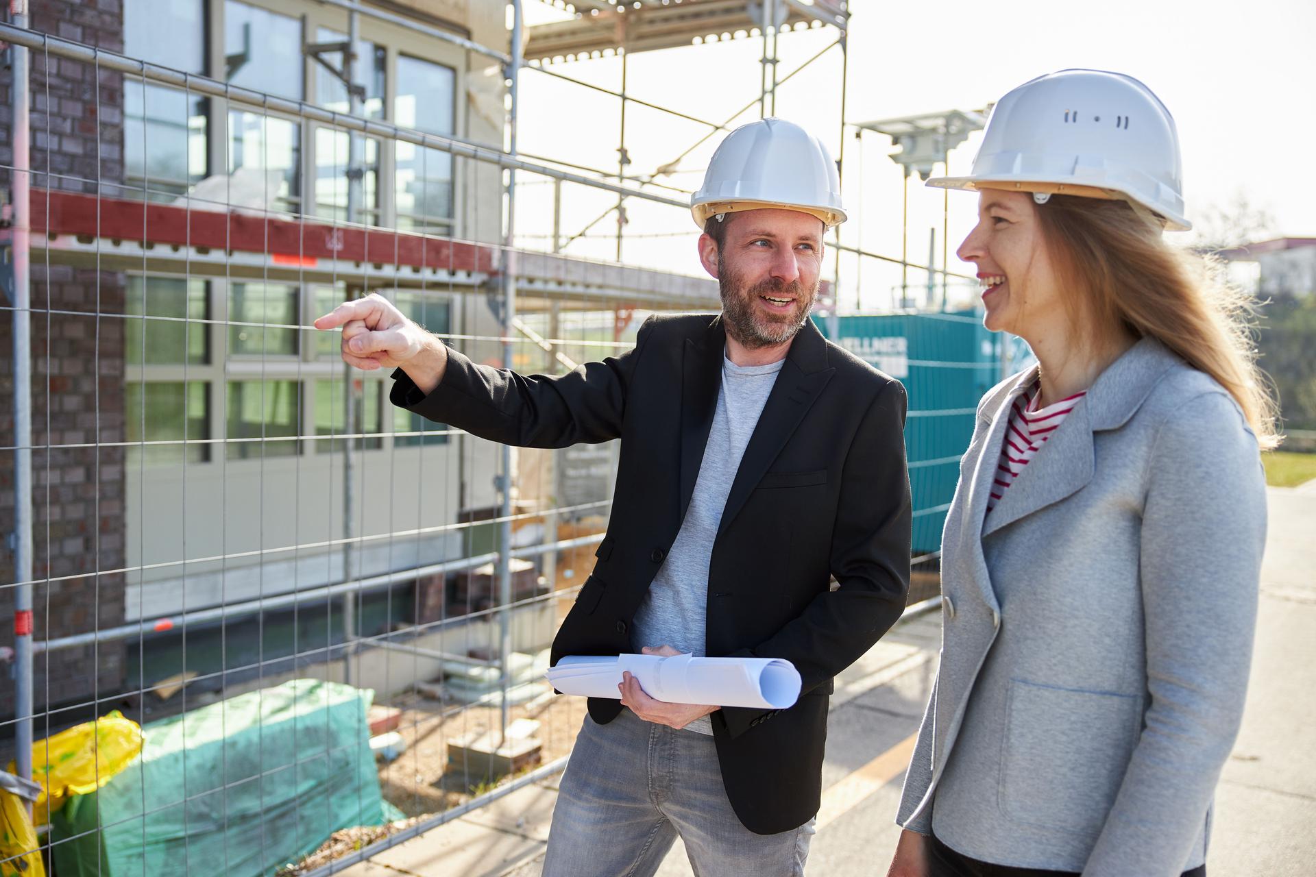 Ein Architekt oder Bauingenieur mit Planungsunterlagen zeigt einem Kunden etwas vor einer Baustelle und beide tragen weiße Harthüte